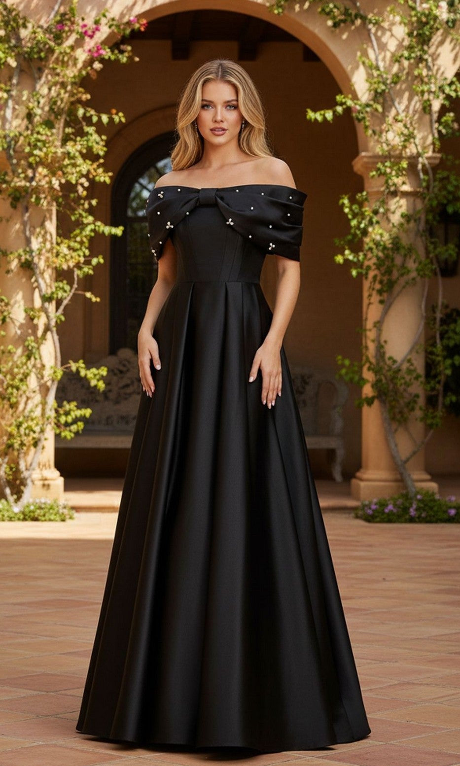 A woman stands outdoors in the Blondie Nites 6154BN long black prom dress, featuring off-the-shoulder styling and jewel embellishments, set against a stone building and greenery.